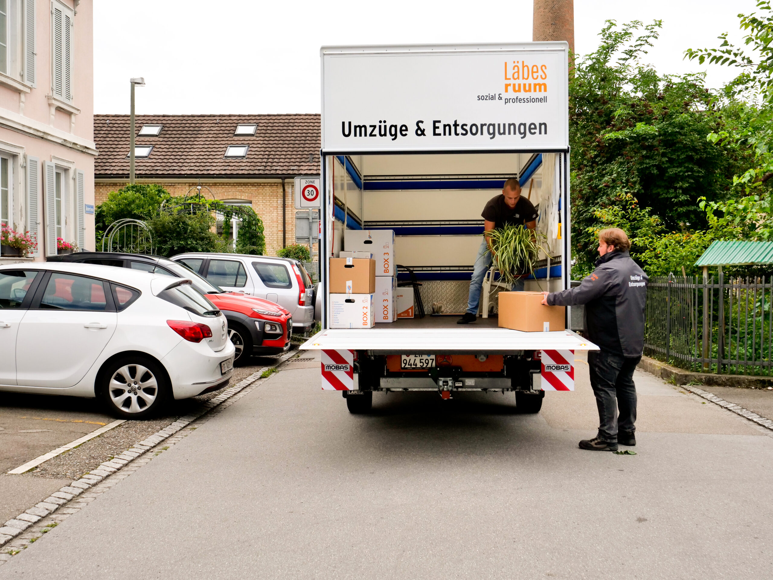 Zwei Mitarbeiter des Läbesruums tragen Pflanzen in einen Umzugslastwagen.