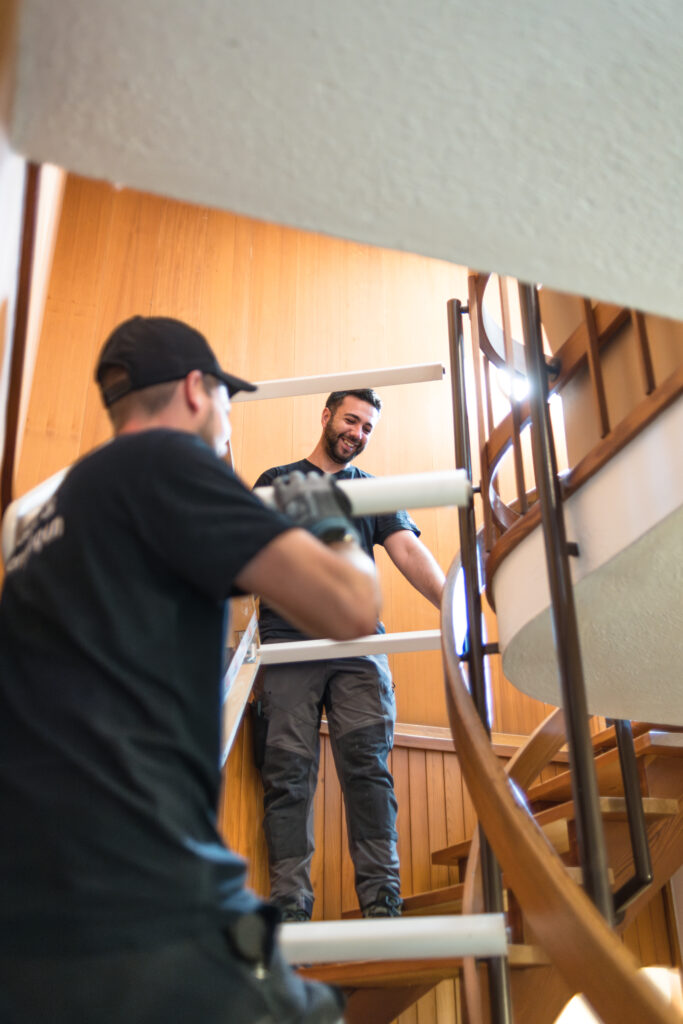 Zwei Mitarbeiter des Läbesruums tragen einen Tisch eine Treppe hoch.